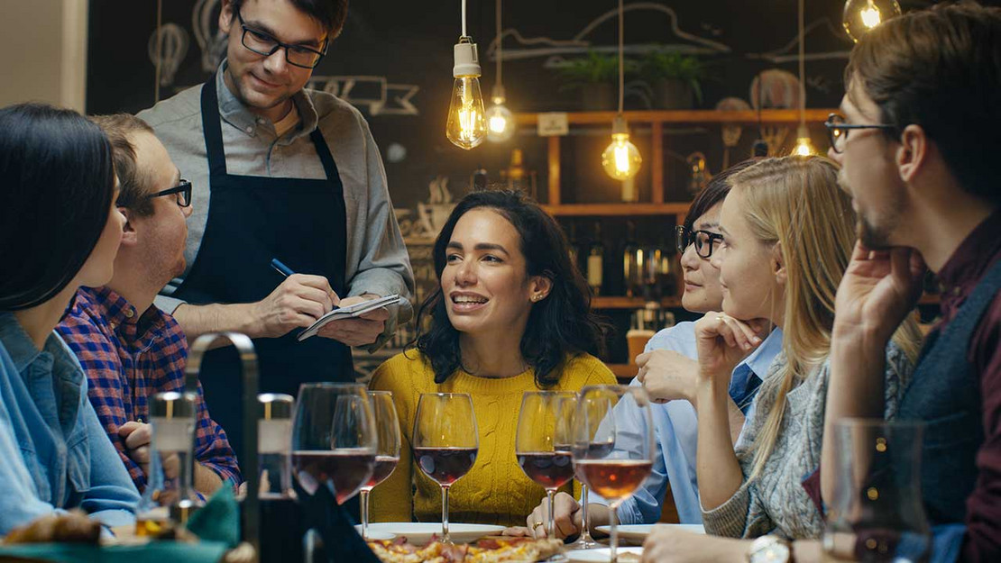 Mehrere Menschen sitzen mit Weingläsern an einem Tisch im Restaurant. 