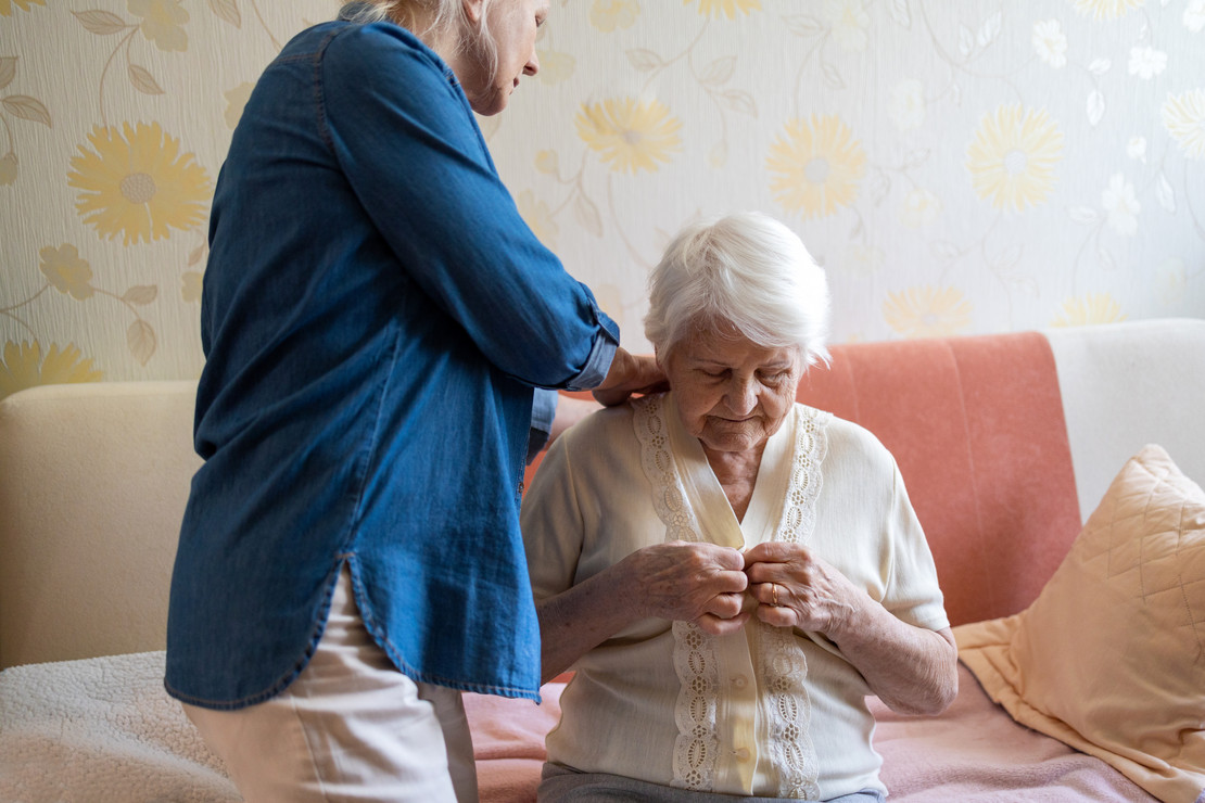 Eine stehende Frau hilft einer sitzenden alten Frau beim Anziehen einer Bluse