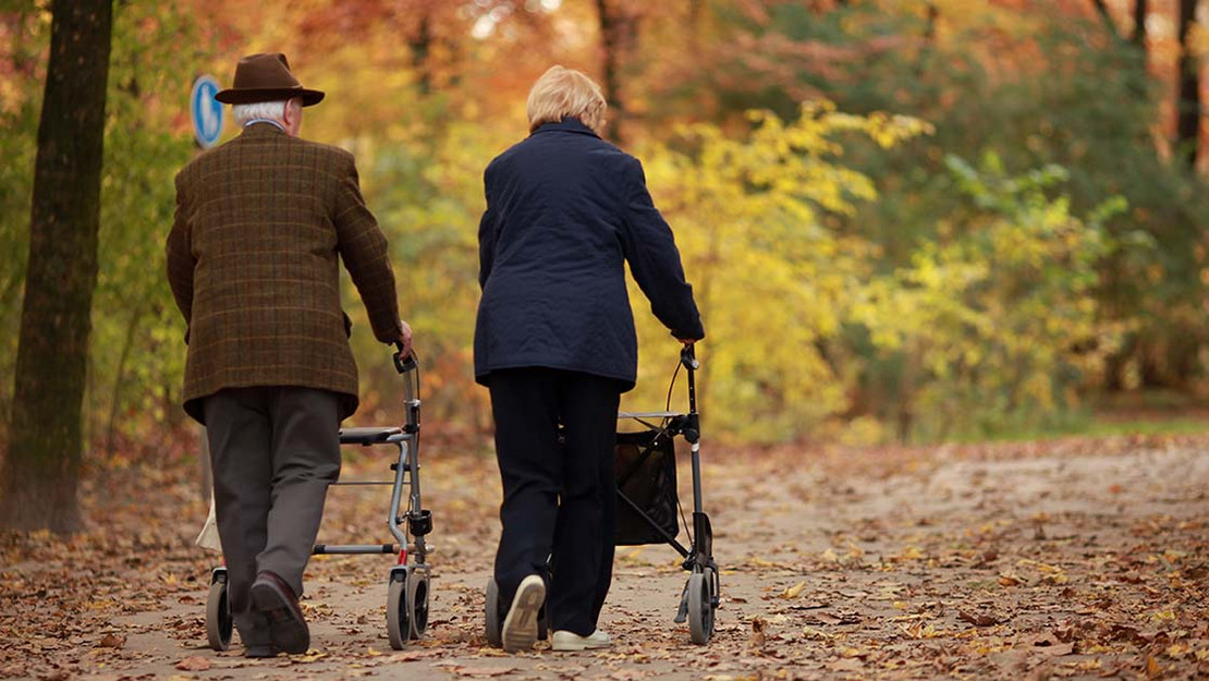 Zwei ältere Personen mit Rollatoren laufen auf einem laubbedeckten Waldweg. 