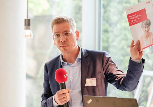 Mann steht an einem Pult und hält die SoVD-Broschüre "Pflegetagebuch" in die Luft. 