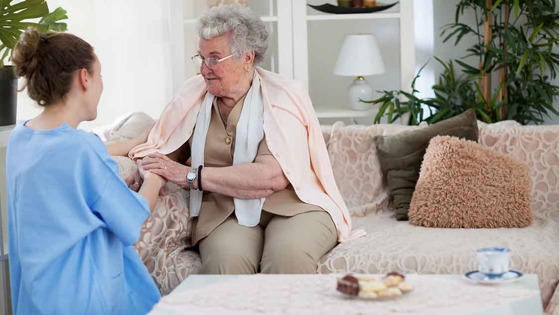 Pflegekraft sitzt einer älteren Frau gegenüber. 